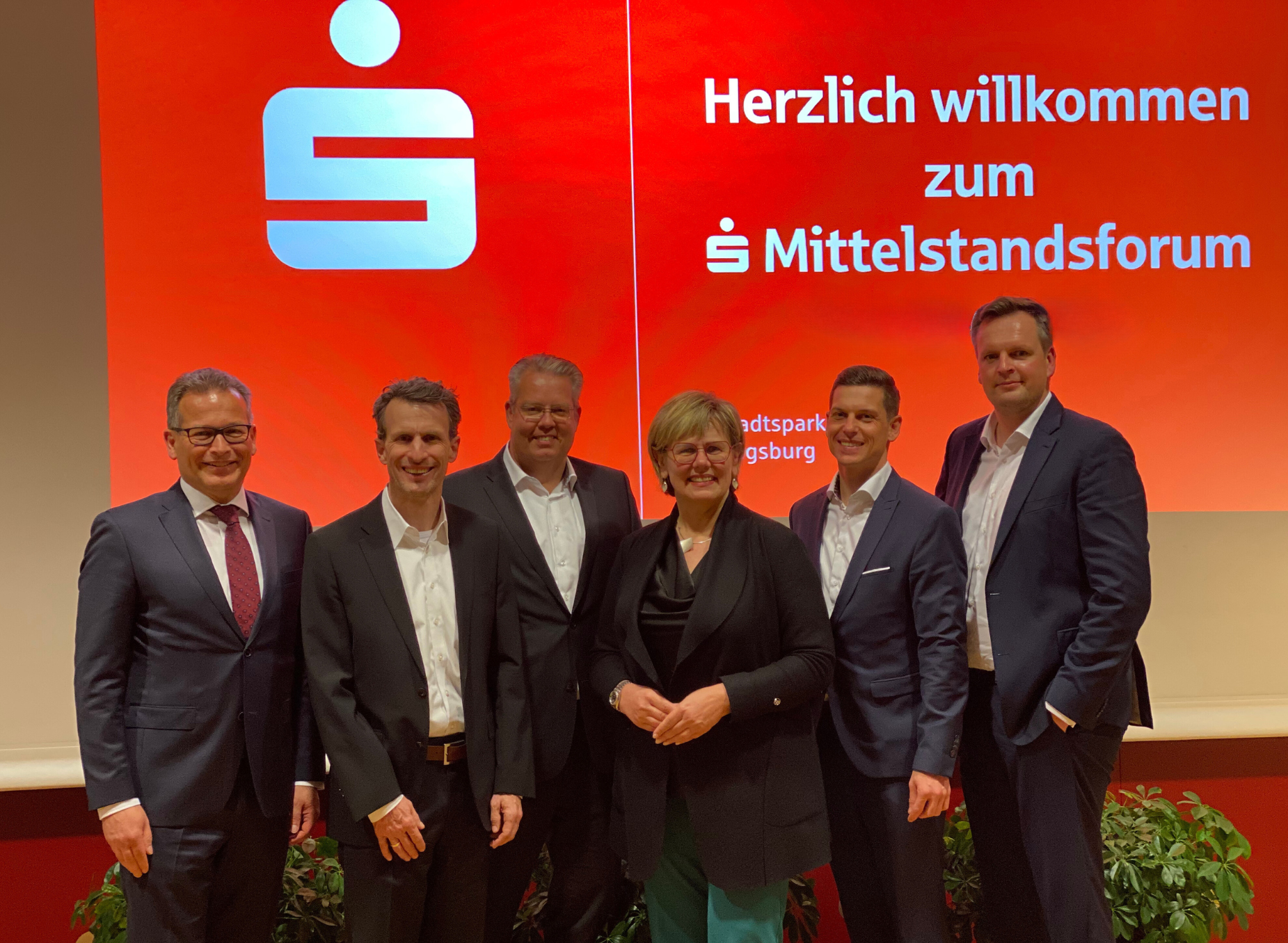 Wir blicken auf eine Gruppe von sechs Personen, fünf Herren und einer Dame, die Businessoutfits tragen. In ihrem Hintergrund ist eine Leinwand zu sehen, auf die vor rotem Hintergrund "Herzlich willkommen zum Sparkassen- Mittelstandsforum" projiziert wird.
