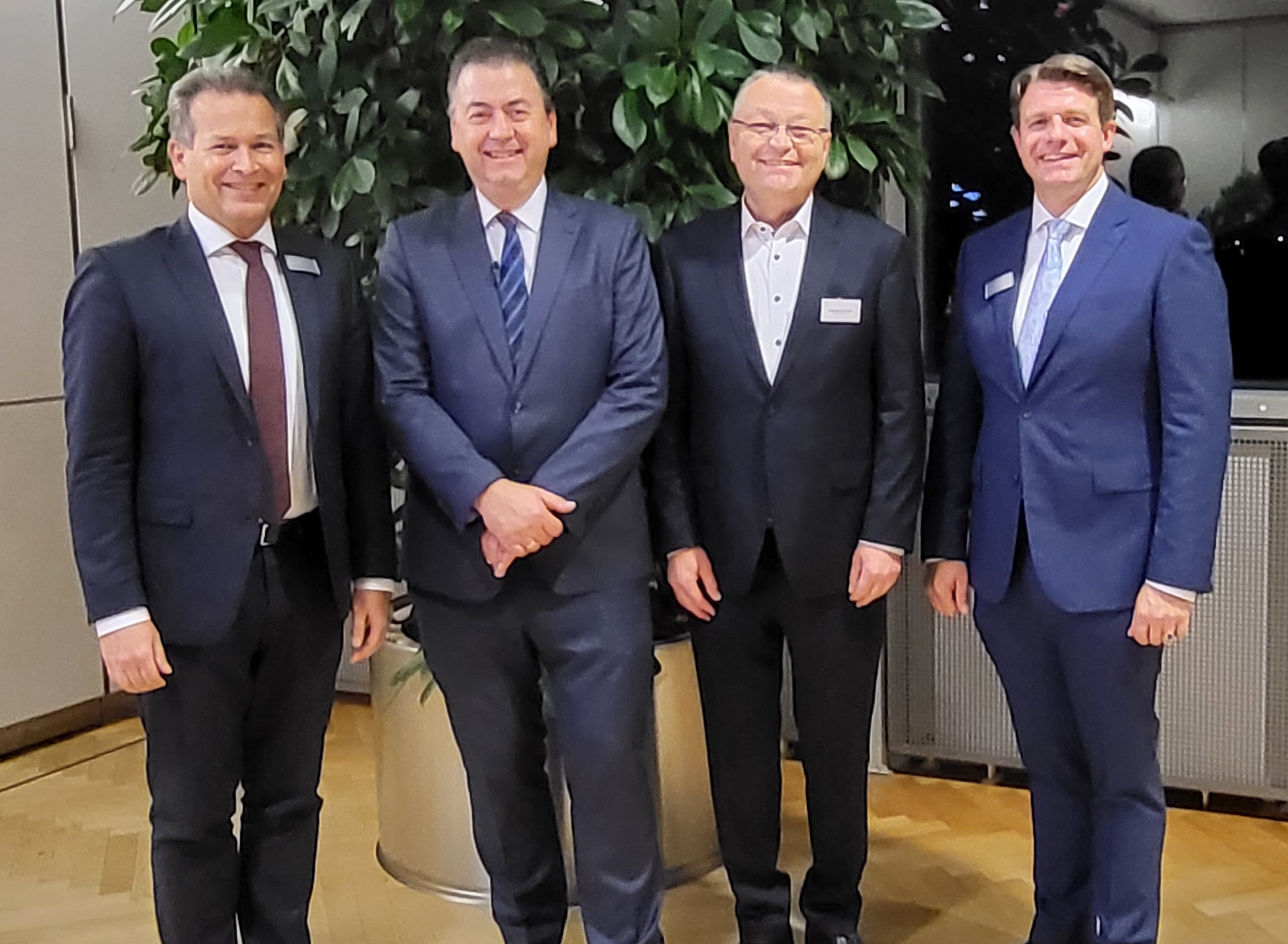 Eine Gruppe von vier Herren in Businessanzügen steht in der Ecke eines Geschäftsraumes vor einer großen Büropflanze. Die Herren lächeln in die Kamera.