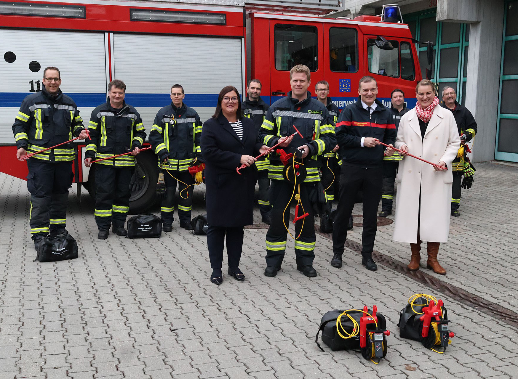 Wir blicken im Innenhof einer Feuerwehrwache auf eine großes Einsatzfahrzeug. In dessen Vordergrund steht eine Reihe von Feuerwehrmännern, die Ausrüstungsgegenstände halten. Im Bildvordergrund sehen wir vier Personen. Zur linken eine elegant gekleidete dunkelhaarige Dame, die einem Herrn in Feuerwehruniform an ihrer rechten Seite einen Ausrüstungsgegenstand reicht. Daneben ein weiterer Herr in einer Funktionsjacke der Feuerwehr. Er bekommt von einer weiteren, elegant gekleideten Dame zu seiner rechten einen Ausrüstungsgegenstand gereicht.