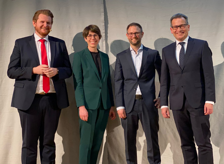 Wir sehen eine Gruppe von vier lächelnden Menschen leicht erhöht auf einer Bühne vor einem weißen Vorhang stehen. Alle Personen sind mittleren ALters. links steht ein Herr im dunklen Anzug mit roter Krawatte, rechts von ihm eine Dame im grünen Kostüm, neben ihr zwei Herren im dunklen Anzug. Alle Personen schauen leicht nach unten direkt in die Kamera