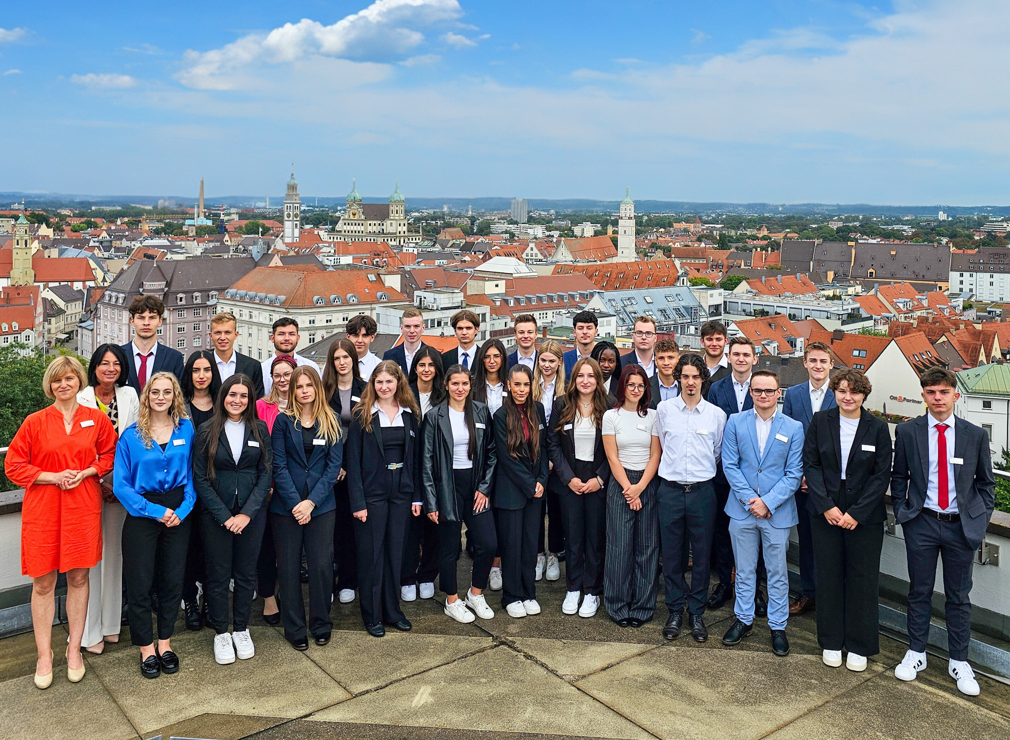 EIne Gruppe von jungen Menschen in Business Casual Kleidung steht auf einer Dachterrasse. Im Bildhintergrund sind die Dächer Augsburg zu sehen. AM linken Rand der Gruppe stehen zwei Damen in Businesskleidung. Ale Personen tragen ein einheitliches Namensschild am Revers.