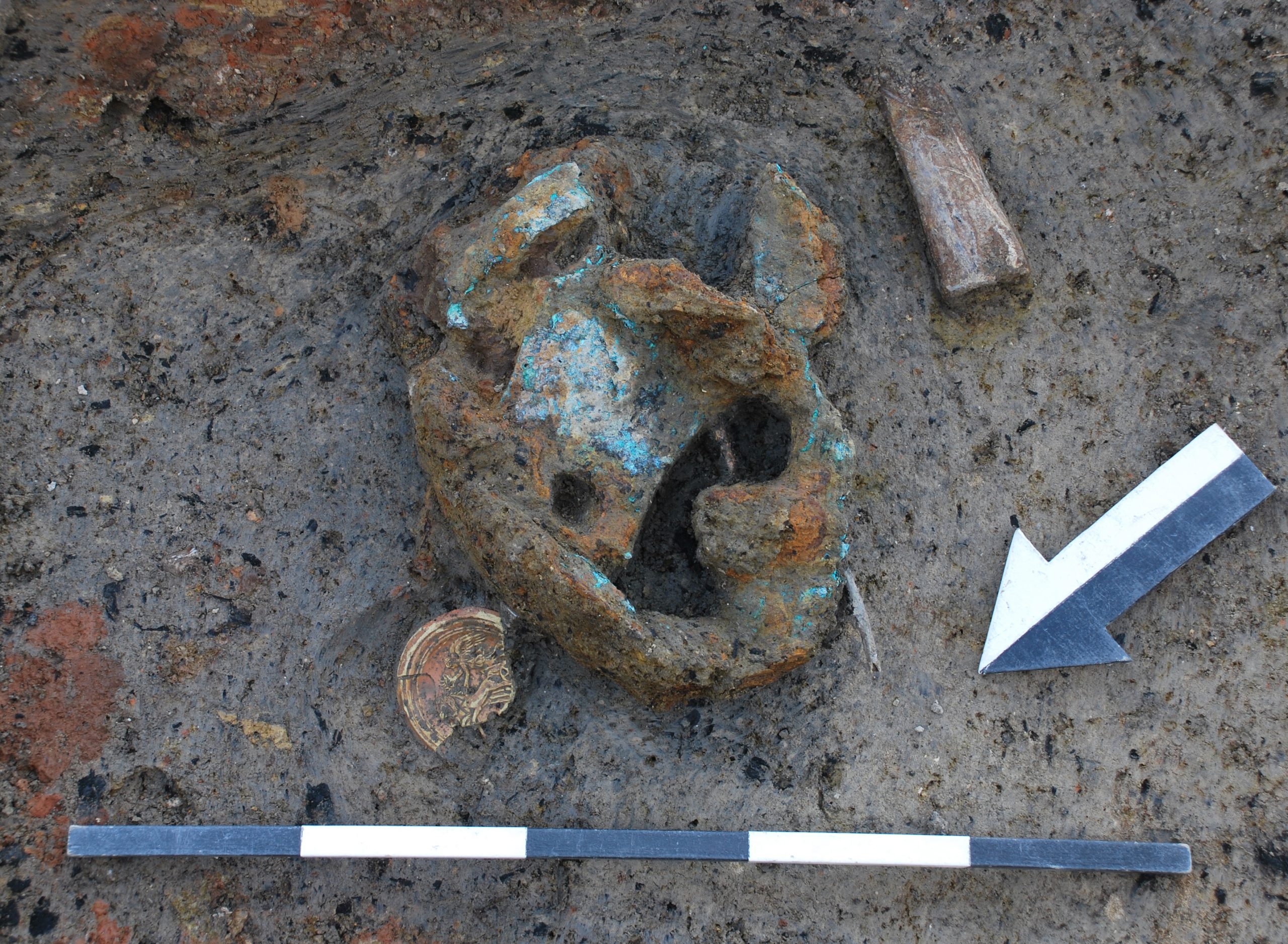 Wir schauen aus der Vogelperspektive aus ca. einem Meter ABstand auf den Boden einer Ausgrabungsstätte. Über einem Meterstab zur Größeneinordnung ragt ein historisches Artefakt aus dem Erdreich. Seine Farbe ist stellenweise grün, stellenweise rostig, stellenweise dunkelgrau. Das Artefakt ist rund und hat innen ein Loch.