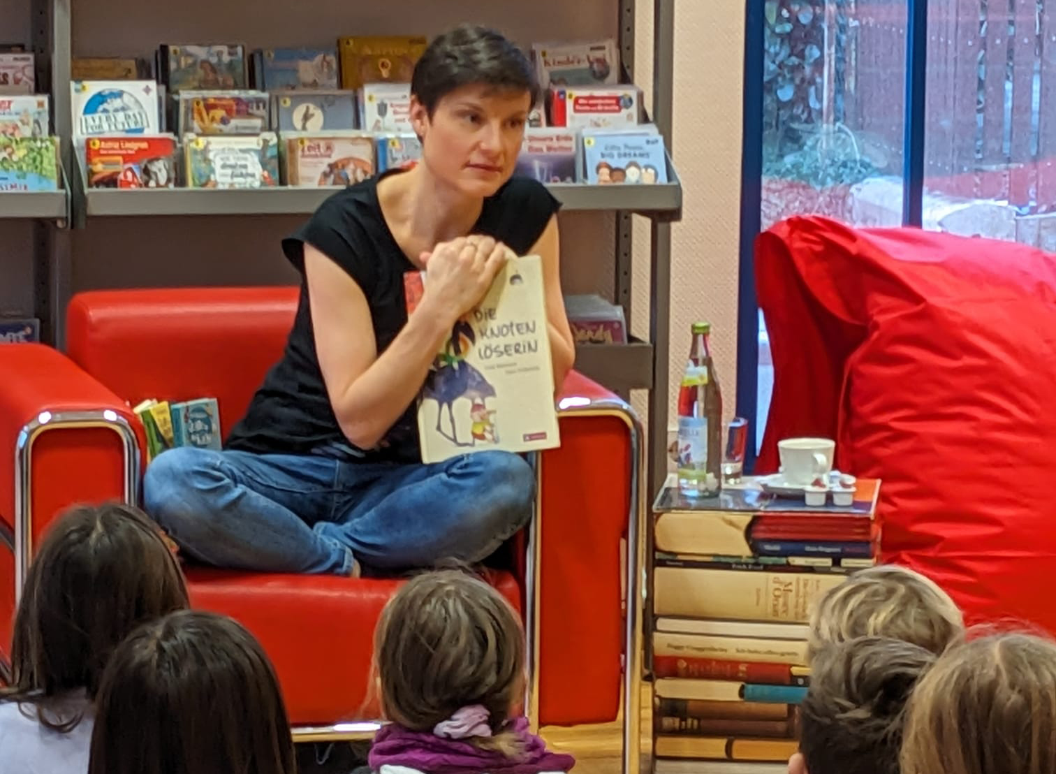 Wir sehen in einen Buchladen. Vor einem Bücherregal ist eine rote Couch aufgebaut. Auf der Couch sitzt eine Dame im Schneidersitz. Sie stützt sich auf ein großformatiges Kinderbuch mit dem Titel "die Knotenlöserin". Sie blickt konzentriert ins Publikum vor ihr: eine Gruppe Kinder, die von hinten zu sehen sind.