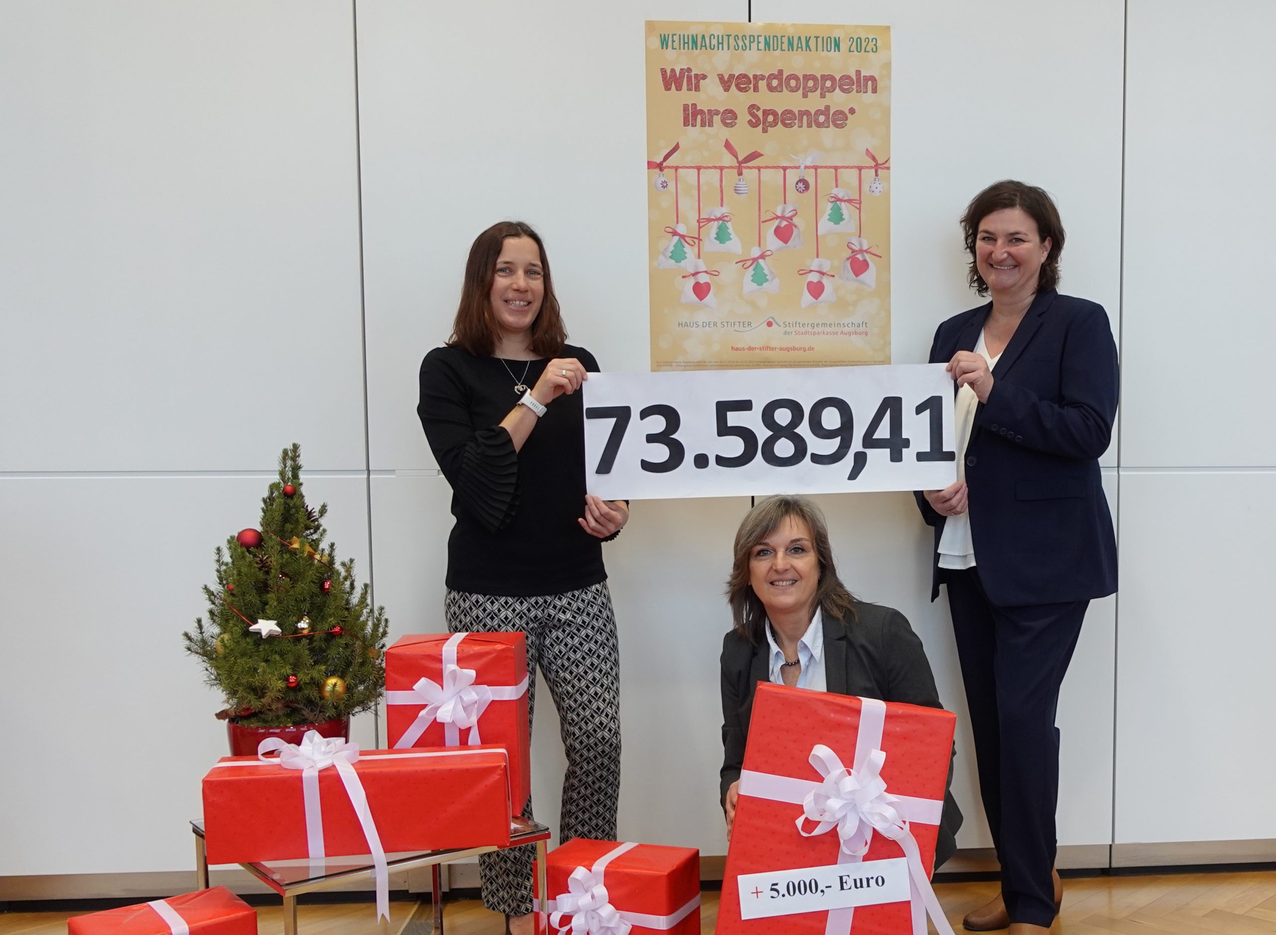 Zu sehen ist eine Gruppe von drei Frauen vor einer weißen Bürowand. An dieser Wand ist ein Plakat, das die Spenden-Verdoppelungsaktion der Sparkasse bewirbt, angebracht. Auf dem Boden des Büros stehen ein kleiner, geschmückter Weihnachtsbaum und viele rot verpackte Geschenkboxen mit weißen Schleifen. die zwei außenstehenden Damen halten eine weiße Banderole, auf der in großen, schwarzen Zahlen "73.589,41" steht.