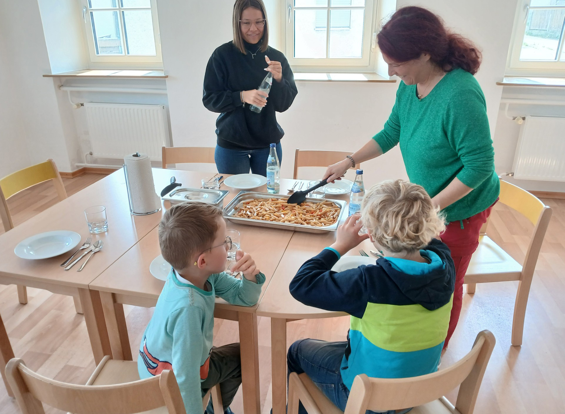 Wir sehen in einen Raum, in dessen Mitte ein gedeckter Tisch steht. An dem Tisch sitzen mit dem Rücken zur Kamera zwei ca. achtjährige Kinder. Um den Tisch herum stehen zwei Damen in legerer Alltagsbekleidung. Eine Dame schraubt gerade eine Glasflasche mit Mineralwasser auf, die andere setzt dazu an, mit einem Servierlöffel Nudelauflauf aus einer Auflaufform, die in der Mitte des Tischs steht, zu schöpfen.