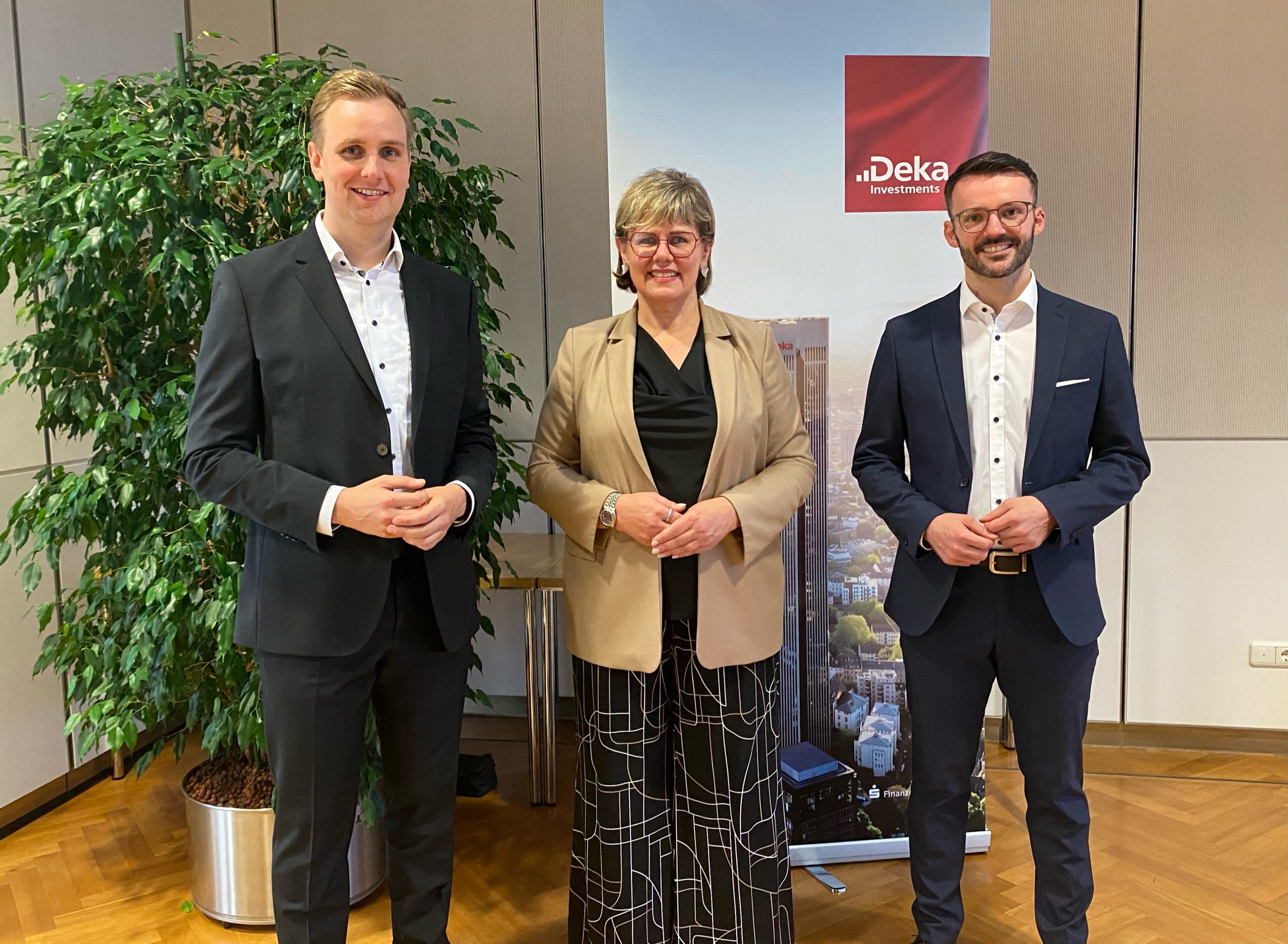 Wir sehen auf eine Gruppe von drei Menschen, die in der Ecke eines Geschäftsgebäudes stehen. Zwei Herren flankieren eine Dame. Alle tragen Businessoutfits. In ihrem Hintergrund stehen eine große Büropflanze und ein Rollup mit der Skyline einer Stadt und einem Deka Investments- Logo