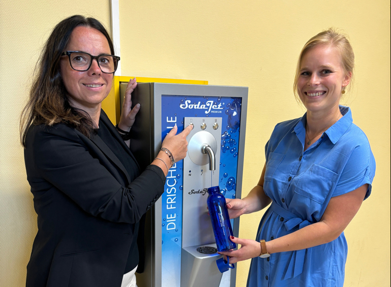 Zwei Damen stehen neben einem Wasserautomaten. SIe tragen sommerlich- elegantere Freizeitkleidung. Eine Dame hält eine dunkelblaue Trinkflasche unter die Wasserausgabe des Automaten. Beide Damen lächeln freundlich in die Kamera.