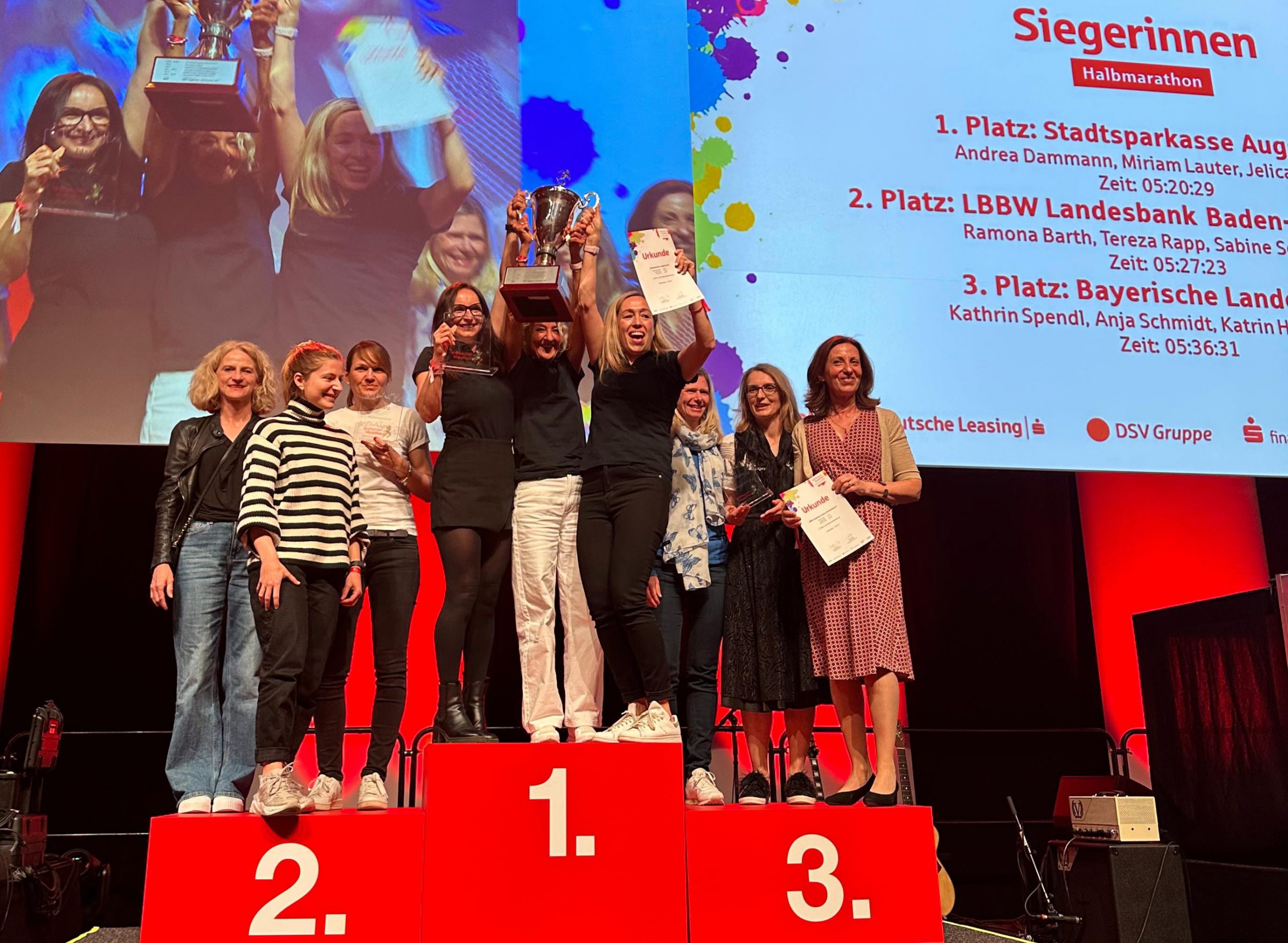 Wir blicken auf eine stimmungsvoll beleuchtete Bühne, auf der gerade eine Preisverleihung stattfindet. Auf einem roten Siegerpodest stehen auf dem 1., 2., und 3. Platz jeweils drei Frauen. Das Trio auf dem ersten Platz in der Mitte reckt jubelnd einen großen, goldenen Pokal und eine Urkunde in die Luft. Auf einer am Bühnenhintergrund befestigten Leinwand ist auszugsweise zu lesen: Siegerinnen Halbmarathon: 1. Platz: Stadtsparkasse Augsburg, 2. Platz LBBW Landesbank Baden-Württemberg, 3. Platz: Bayerische Landesbank