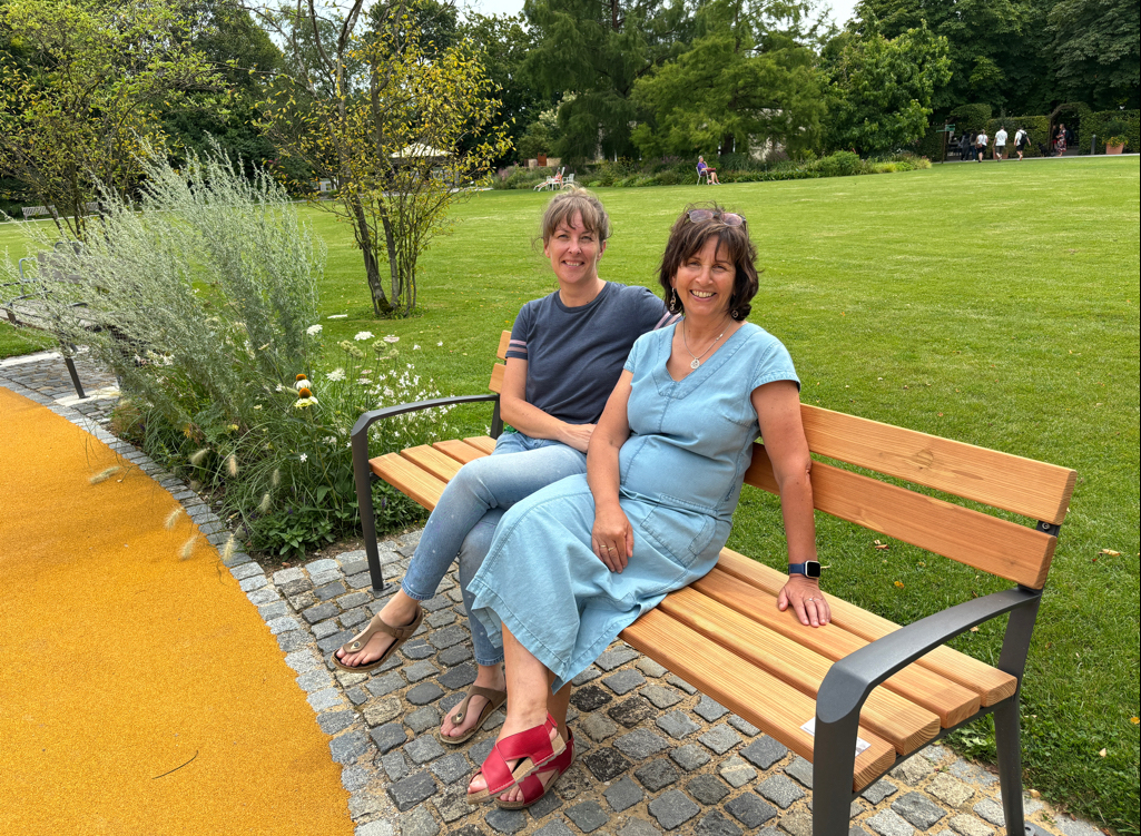 Zwei Damen in sommerlicher Tageskleidung sitzen auf einer Parkbank. Die Parkbank steht zwischen einer Wiese und einem gepflasterten Platz und sieht robust und neu aus und besteht aus hellem Holz und dunklem Metall. Die Damen lächeln freundlich in die Kamera.