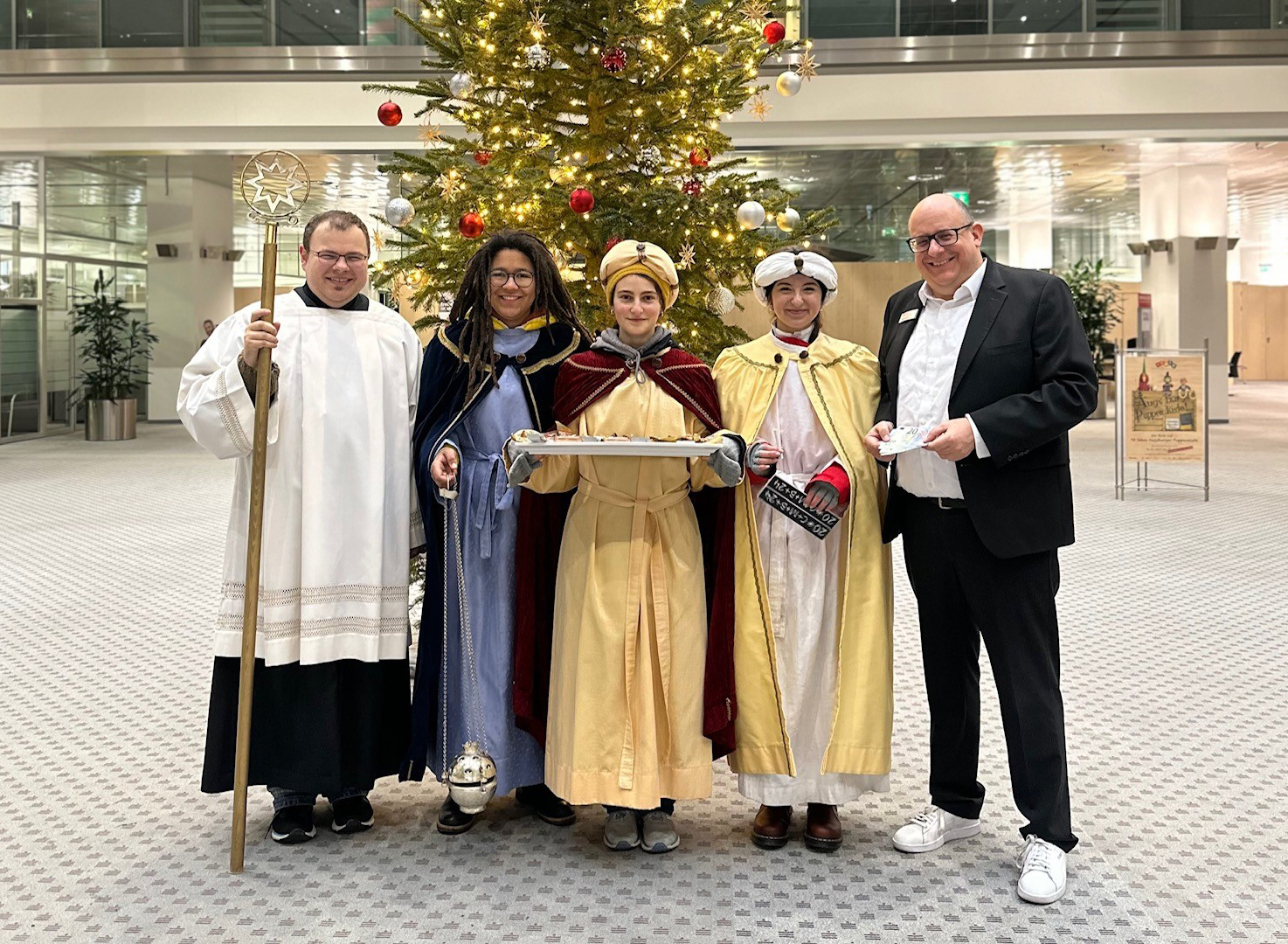 Zu sehen sind fünf Personen, die im Foyer einer großen Bankfiliale vor einem Weihnachtsbaum stehen. In der Mitte drei ca. 12 jährige Kinder, die als heilige drei Könige verkleidet sind. Links von Ihnen ein Bischof im Ornat, rechts der Leiter der Bankhauptstelle in Anzug und Sneakern. Der Bankangestellte hält einige Geldscheine in der Hand und ist im Begriff, diese zu überreichen.