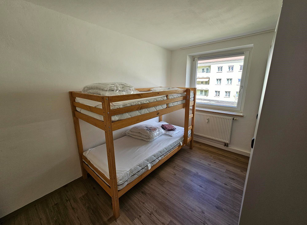 Wir blicken auf ein Stockbett in der Ecke eines kleinen Zimmers. Auf dem Bett liegen zwei neue Matratzen und zwei neu verpackte Sets mit Bettwäsche. Durch ein Fenster im Raum scheint Sonne herein. Außerdem gibt das fenster den Blick auf ein gegenüberliegendes Mehrfamilienhaus frei.