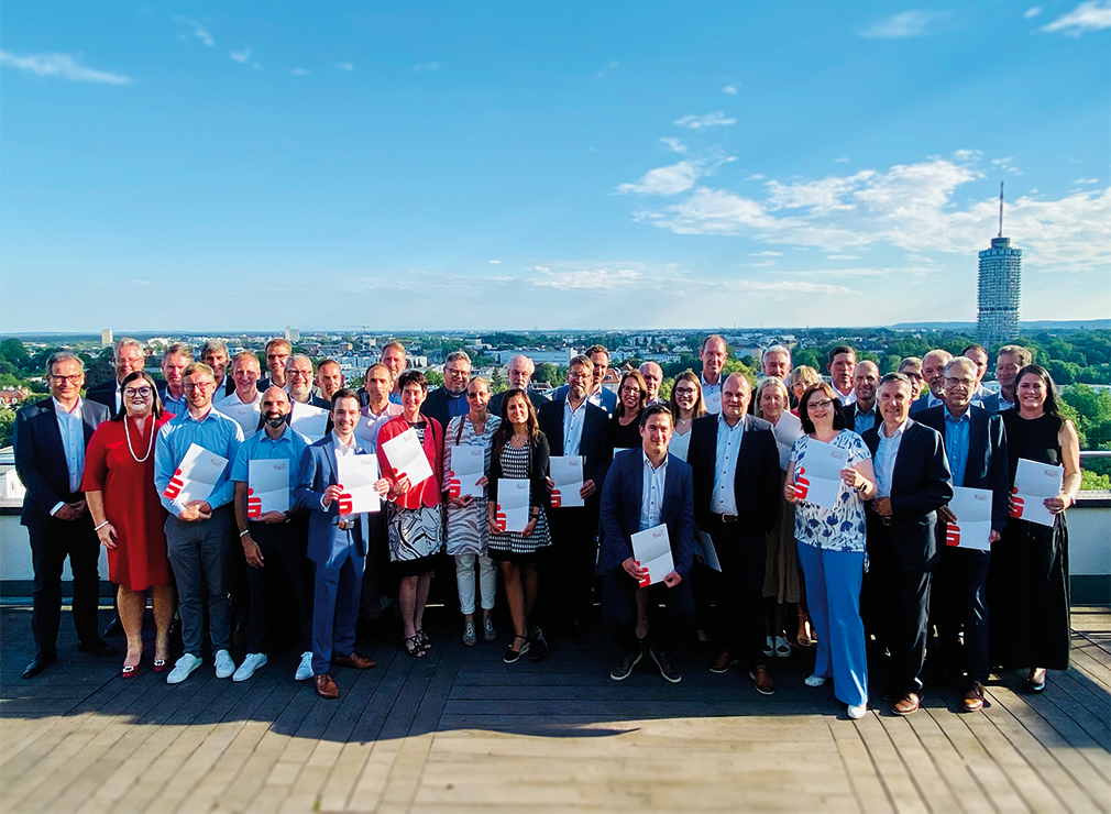 Eine große Gruppe von Menschen in Businesskleidung steht auf einer sonnigen Dachterrasse. Im Hintergrund des Bildes, also um die Dachterrasse herum, ist die Skyline von Augsburg zu sehen. Die Personen halten Zertifikate in der Hand. Alle Personen und die Umgebung sind in helles Sonnenlicht getaucht.