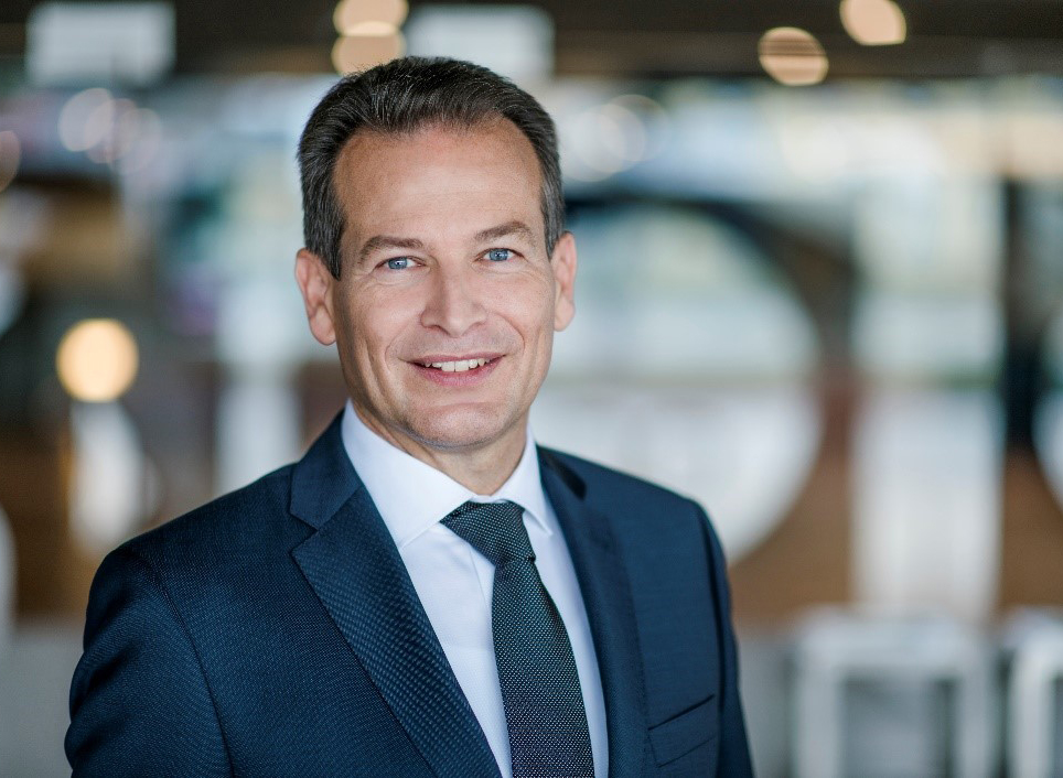 Wir sehen einen Herren im blauen Businessanzug mit passender Krawatte und weißem Hemd. Das Bild ist als Portrait bis zur Brust aufgenommen. Im Hintergrund sieht man in Unschärfe das Innere eines Geschäftsgebäudes. Der Herr lächelt souverän in die Kamera.