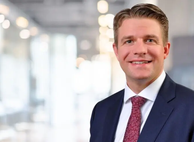 Ein Herr im Businessanzug lächelt freundlich in die Kamera. Im Hintergrund ist in Unschärfe das Innere eines Geschäftsgebäudes zu erkennen.