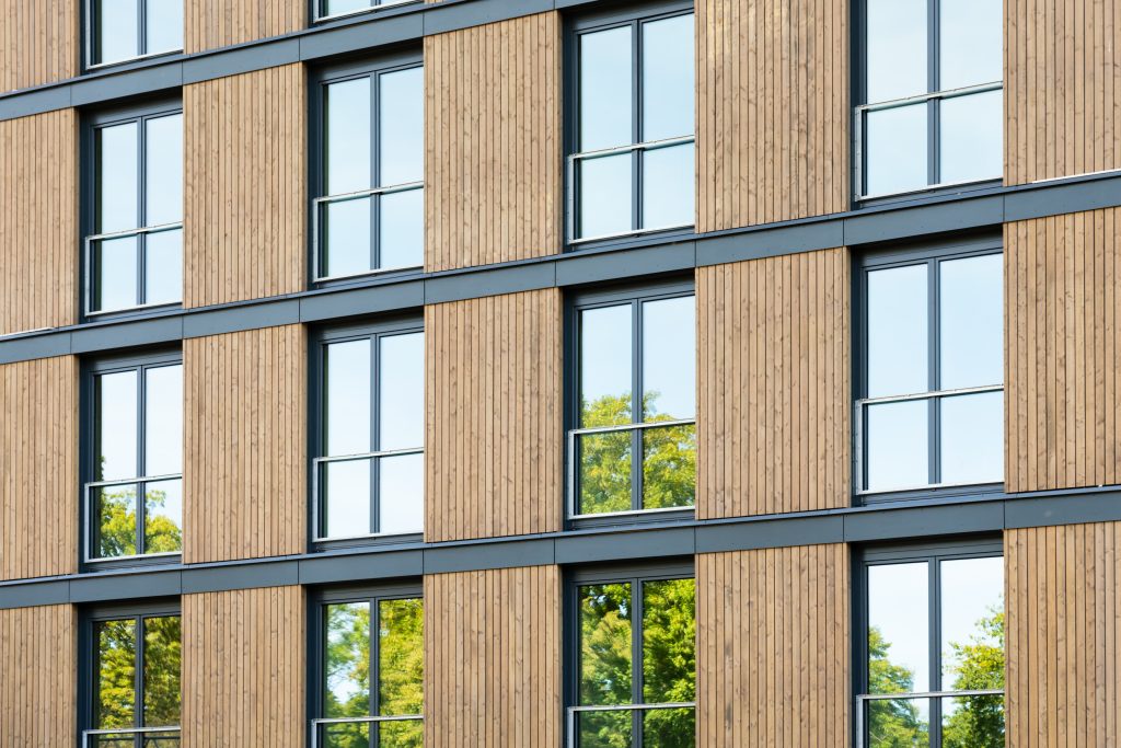 Holzverkleidete Fassade eines modernen Gebäudes. In den Fenstern spiegeln sich Baumkronen.